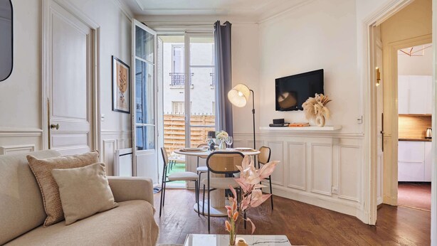 A living room with a sofa, a coffee table with vases, a dining area with chairs and a table, a TV on the wall, and a kitchen visible through an open door. - Eiffel Tower Terrace Apartment (Paris)