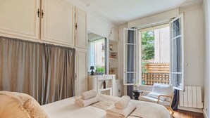 A bedroom with a bed, a chair, a window with curtains, and a view of the outside. - Eiffel Tower Terrace Apartment (Paris)