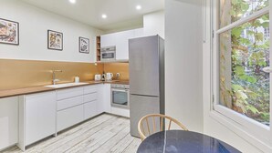 A modern kitchen with white cabinets, a stainless steel refrigerator, a microwave, and a sink. There is a window with a view of greenery outside. - Amazing apartment in the Center of Paris ( Marais) (Paris)