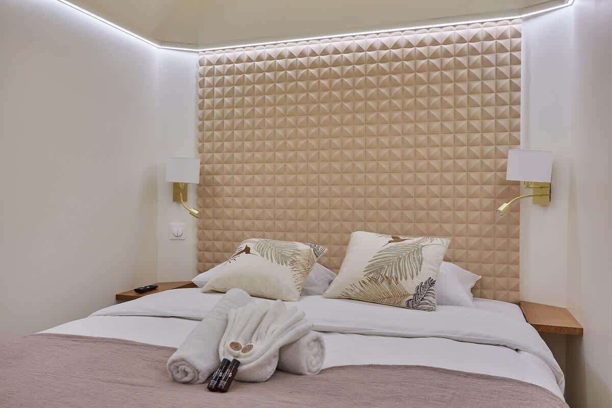 A neatly made bed with pillows and a rolled towel, a wall with a geometric pattern, and two wall-mounted lamps.