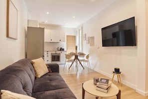 A modern living room with a sofa and a coffee table, a dining area with chairs and a table, a kitchen with cabinets and appliances, and a TV mounted on the wall.