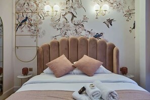 Une chambre à coucher avec un grand lit, deux oreillers roses, une tête de lit en bois et des œuvres d’art murales représentant des oiseaux et des fleurs.