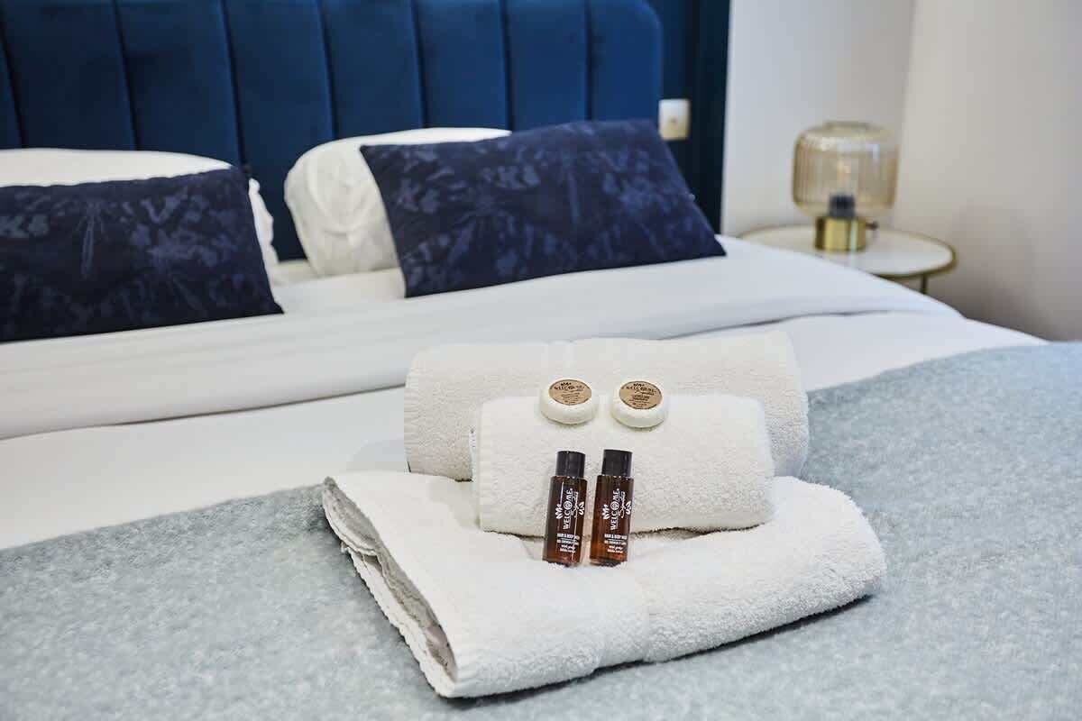 A neatly made bed with a blue headboard, white bedding, and two decorative pillows. A folded towel and two small bottles are placed on the bed.