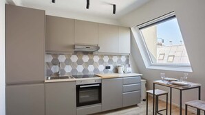 Una cocina moderna con armarios grises, horno integrado, ventana con vistas a edificios y un pequeño comedor con taburetes.