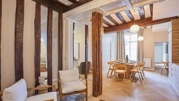 Ein geräumiger Raum mit sichtbaren Holzbalken, einem Essbereich mit Holzmöbeln und einem großen Fenster, das natürliches Licht hereinlässt.
