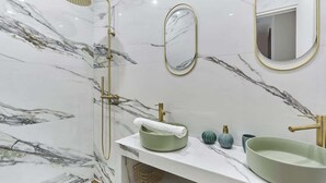 Ein Badezimmer mit Marmorwand, einer freistehenden Badewanne, einem Waschbecken mit grüner Schüssel und einem spiegel mit goldener Rahmung.