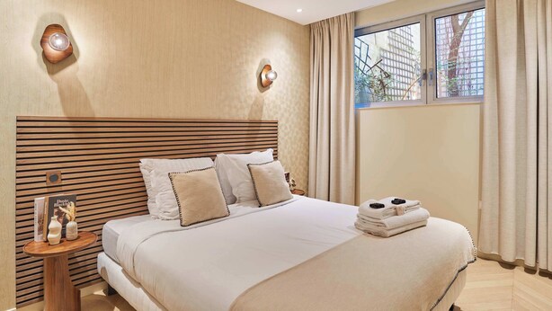 A hotel room with a large bed, wooden headboard, two wall sconces, a window with curtains, and a small table with a book and a vase. - Elegant Apartment St Germain des prés Luxembourg Park (Paris)