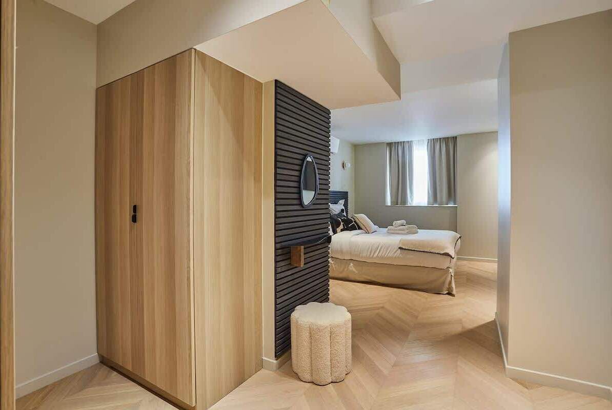 Une chambre à coucher moderne avec une armoire en bois, un petit pouf, un lit recouvert d’une couette blanche et une fenêtre avec des rideaux.