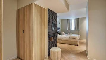 Une chambre à coucher moderne avec une armoire en bois, un petit pouf, un lit recouvert d’une couette blanche et une fenêtre avec des rideaux.