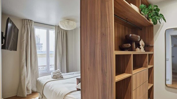 A bedroom with a bed, a window with curtains, a wooden shelving unit, and a potted plant.