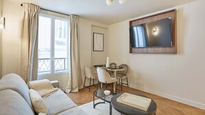 A modern living room with a sofa, a dining table with chairs, a flat-screen TV mounted on the wall, and a large window with curtains. - New apartment Center of Paris/Le Marais (Paris)