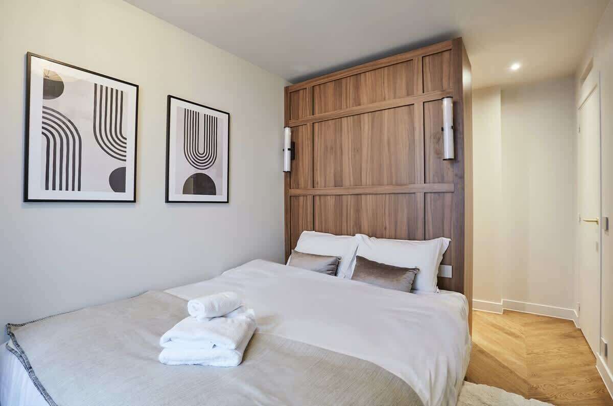 Ein ordentlich bezogenes Bett mit weißen Leinen und einem gefalteten Handtuch, ein hölzerner Kopfteil und zwei gerahmte abstrakte Kunstwerke an der Wand.