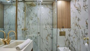 Un baño moderno con ducha envuelta en vidrio, pared de azulejos de mármol, mueble de madera y lavamanos blanco con detalles dorados.