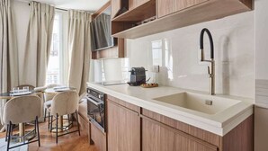A modern kitchen with a white countertop, wooden cabinets, and a stainless steel faucet. There is a dining area with a table and chairs, and a window with curtains. - New apartment Center of Paris/Le Marais (Paris)