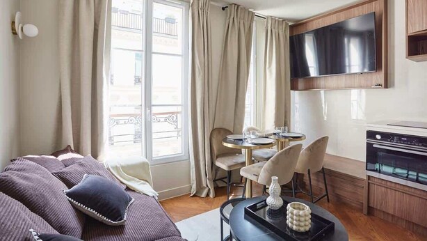 A modern living room with a sofa, a dining area with chairs, a TV mounted on the wall, and a kitchen area with an oven. - New apartment Center of Paris/Le Marais (Paris)