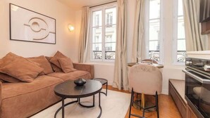 Un salon chaleureux avec un canapé marron, une table basse ronde et une petite table à manger avec une chaise. Il y a une grande fenêtre avec des rideaux, une œuvre d’art accrochée au mur et un coin cuisine avec une cuisinière.