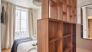 A bedroom with a bed, a wooden wardrobe, a window with curtains, and a wall-mounted mirror.