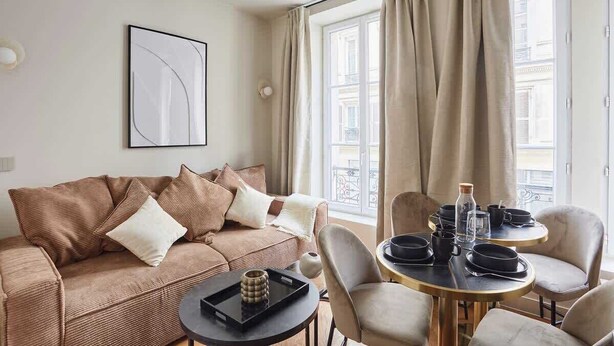 A modern living room with a brown sofa, a round dining table with chairs, and a large window with curtains.