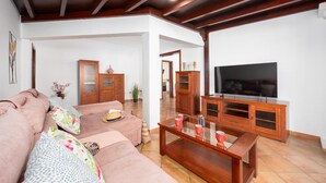 Una sala de estar con un sofá, una mesa de centro y un televisor. Se observan armarios de madera y una planta.