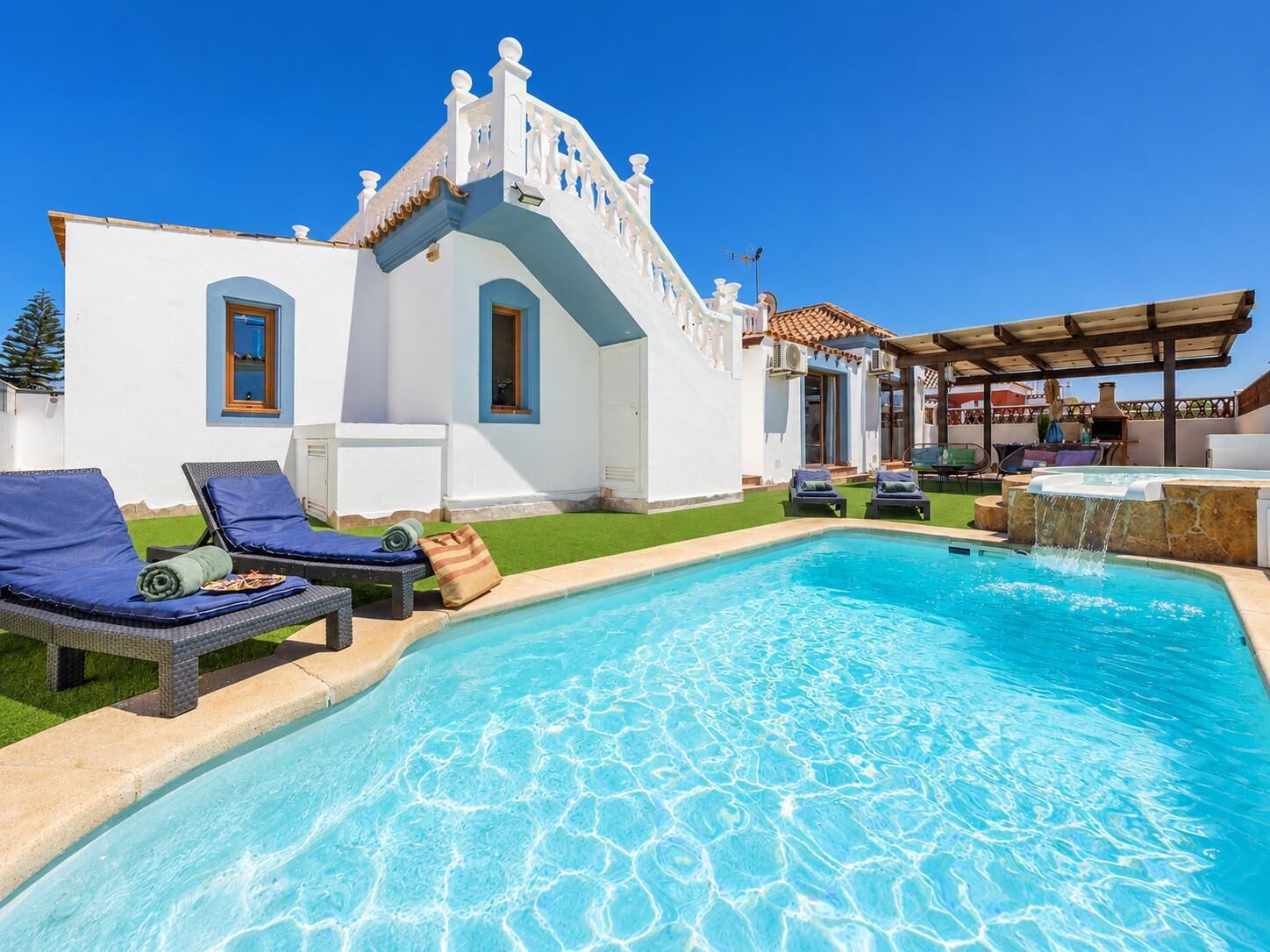Un área de piscina con sillones de descanso, un jacuzzi y un edificio blanco con tejado de baldosas.