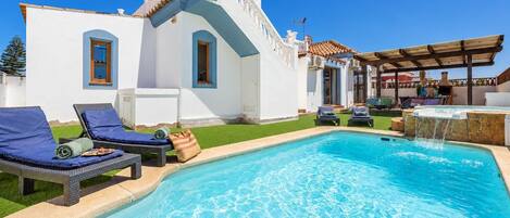 Un área de piscina con sillones de descanso, un jacuzzi y un edificio blanco con tejado de baldosas.