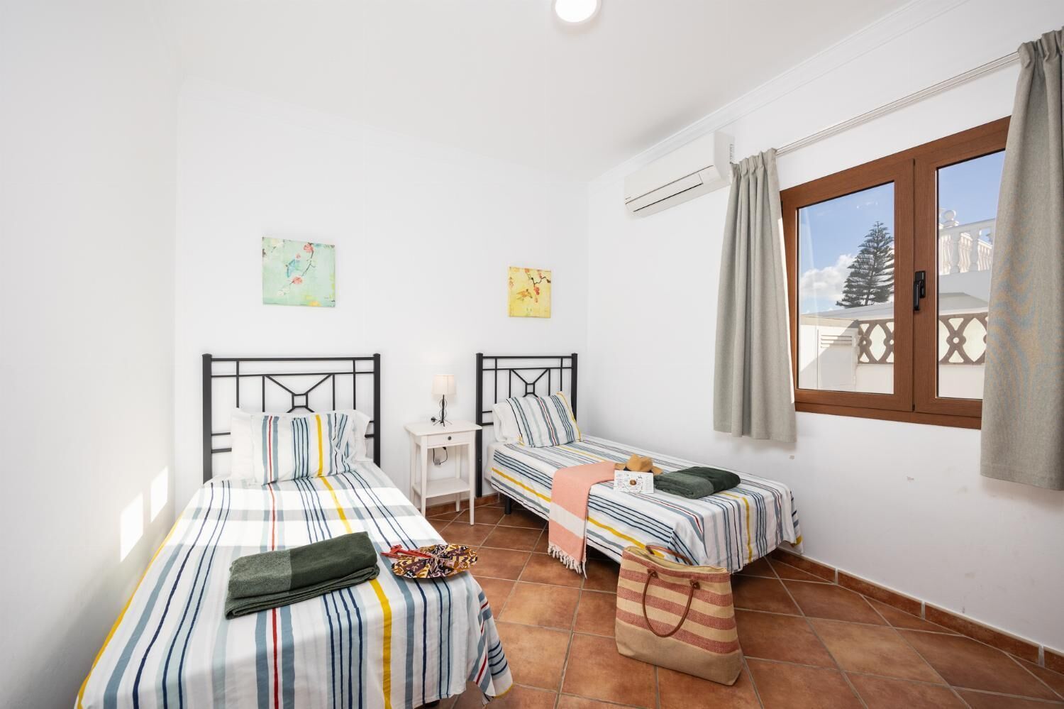 Een slaapkamer met twee eenpersoonsbedden, een klein nachtkastje, een raam met gordijnen en uitzicht op een balkon.