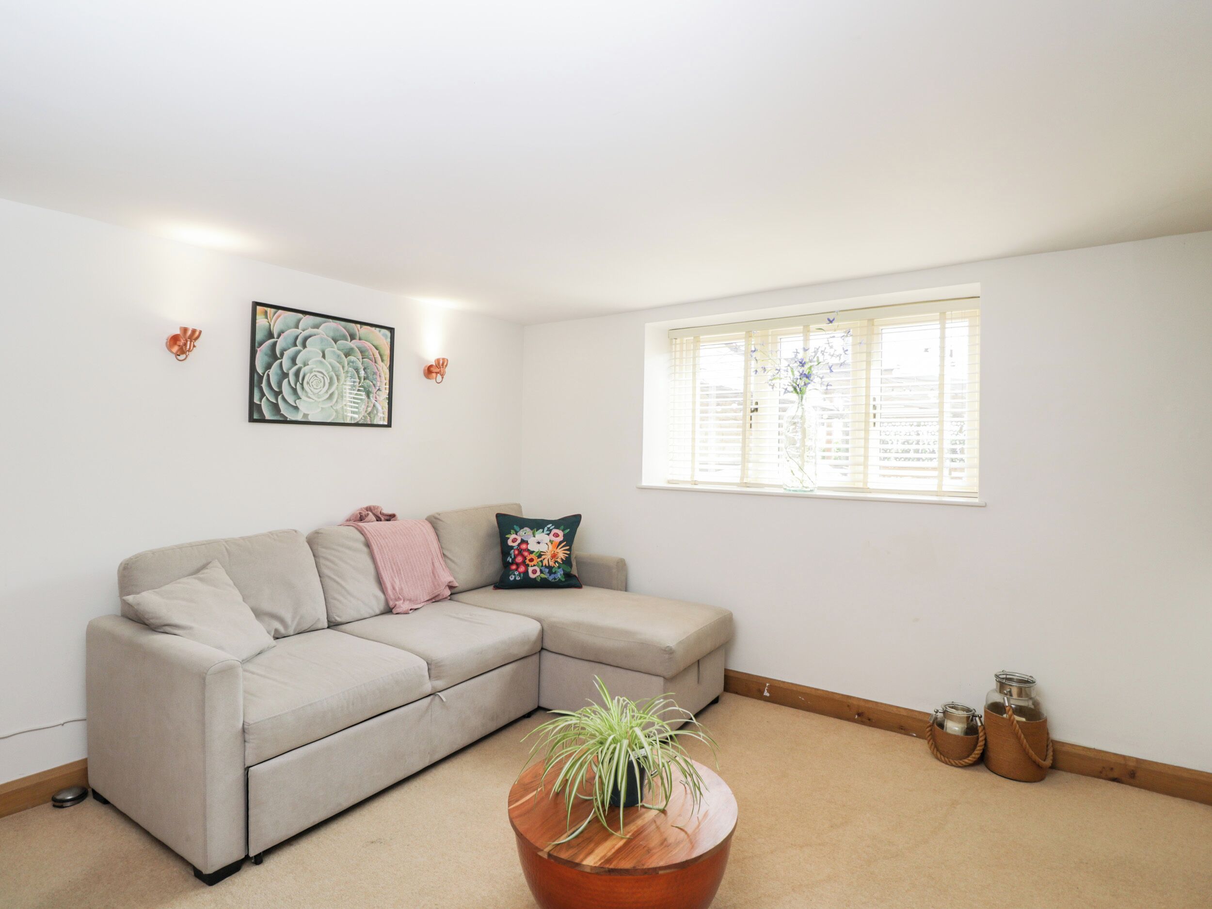 Una sala moderna con un sofá seccional color beige, una mesa de centro redonda de madera con una planta, una obra de arte floral enmarcada y una ventana con persianas.