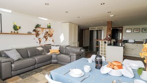 Ein modernes Wohnzimmer mit einem grauen Ecksofa, einem Esstisch, der für ein Frühstück gedeckt ist, und einer Küche im Hintergrund.