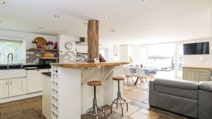 Eine moderne Küche mit Kochinsel, Barhocker, Essbereich mit Tisch und Stühlen sowie ein graues Sofa in einem hellen Wohnraum mit großen Fenstern.