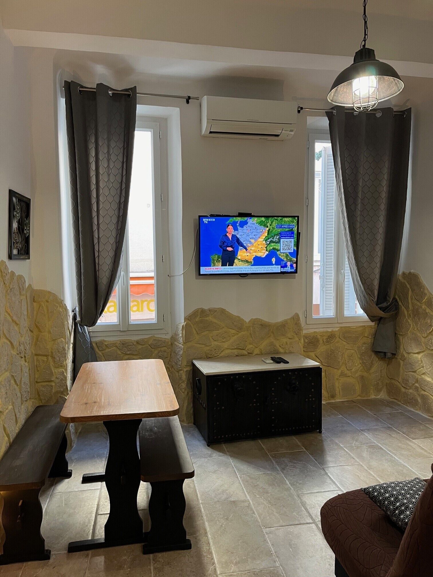 Olohuone, jossa on kiviseinä, puinen pöytä, penkki, sohva, seinälle asennettu televisio ja kivinen takka.