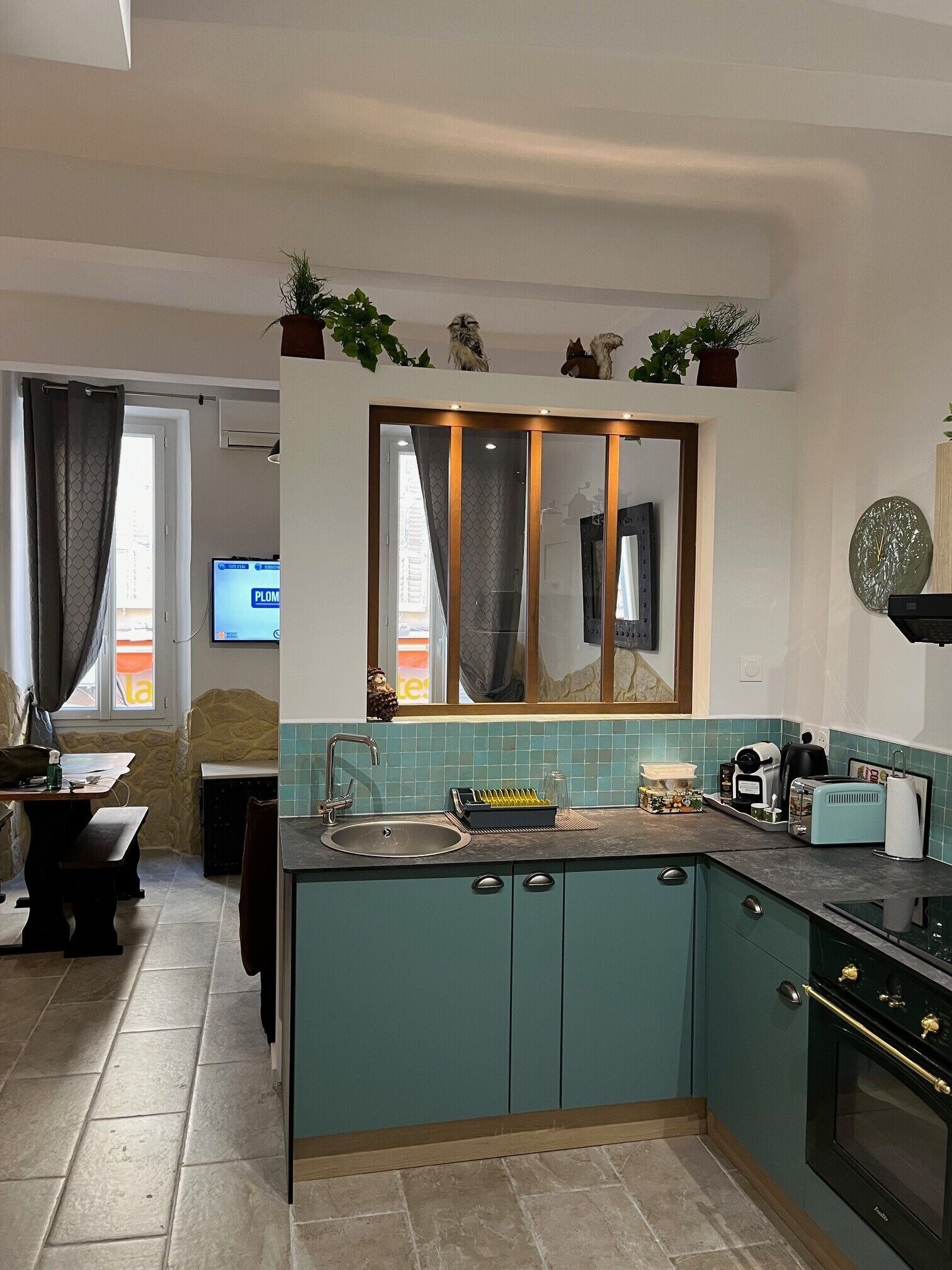 Cocina compacta con armarios color verde azulado, horno empotrado, un lavabo y un ventanal sobre la encimera.