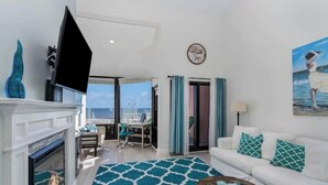 Un salon moderne doté d’une cheminée, d’un grand téléviseur à écran plat, d’un canapé blanc agrémenté de coussins turquoise, d’une table basse et offrant une vue sur l’océan grâce à une grande fenêtre.