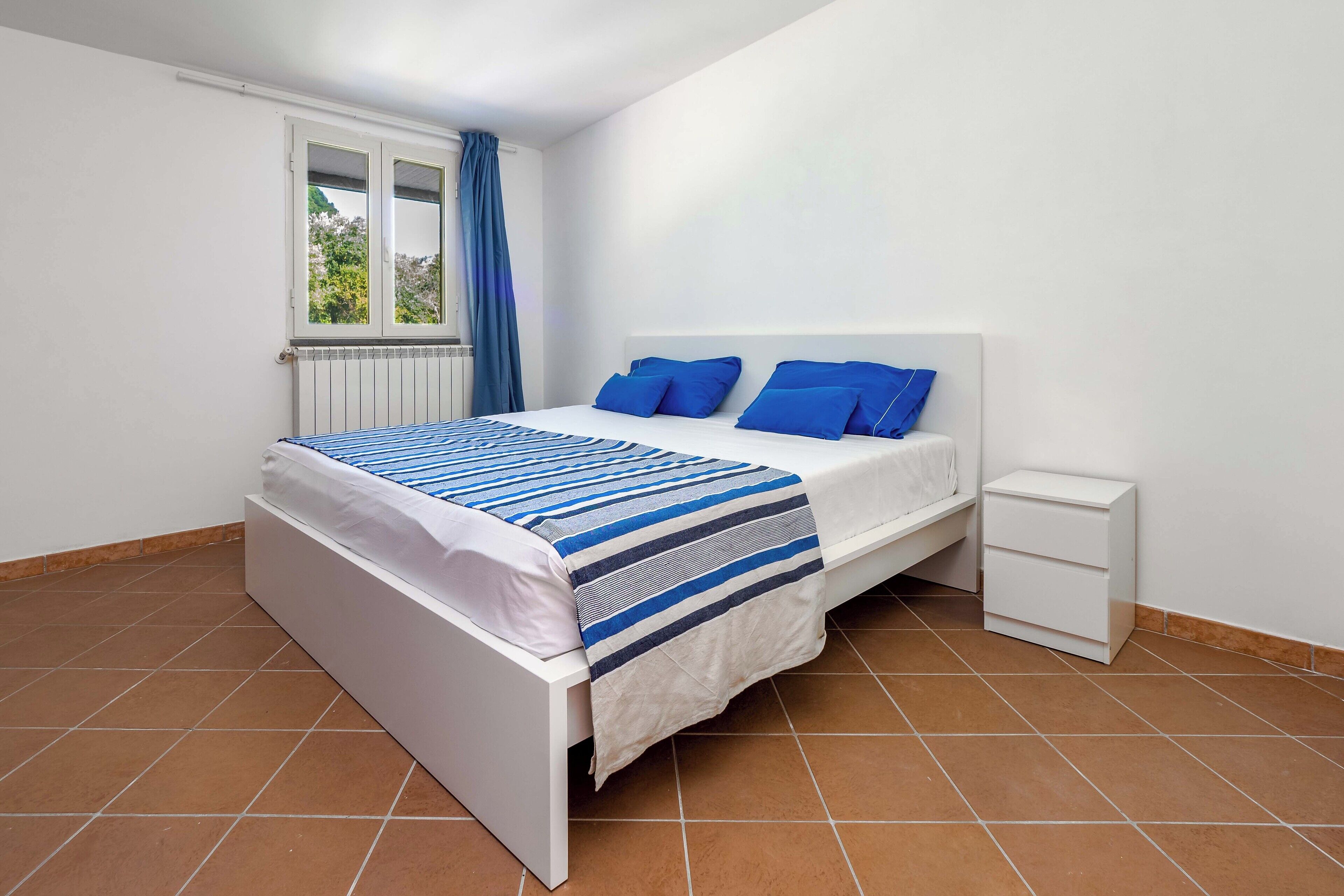 Una camera da letto con un letto bianco, una coperta a righe blu e bianche, un comodino bianco e una finestra con tende blu.