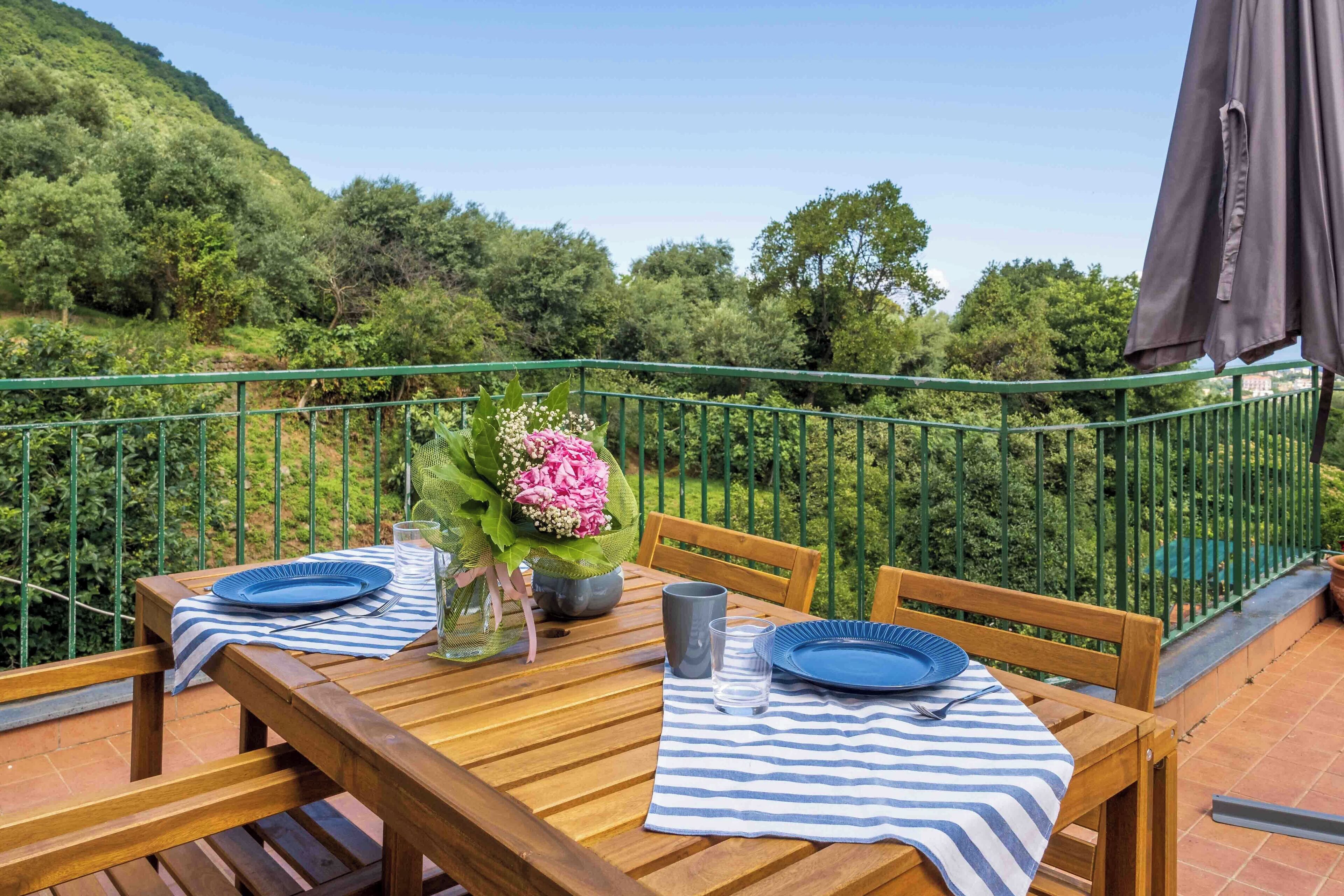 A wooden table set with blue plates, a vase of flowers, and a striped tablecloth on a terrace with a green railing and a view of a forested hillside.