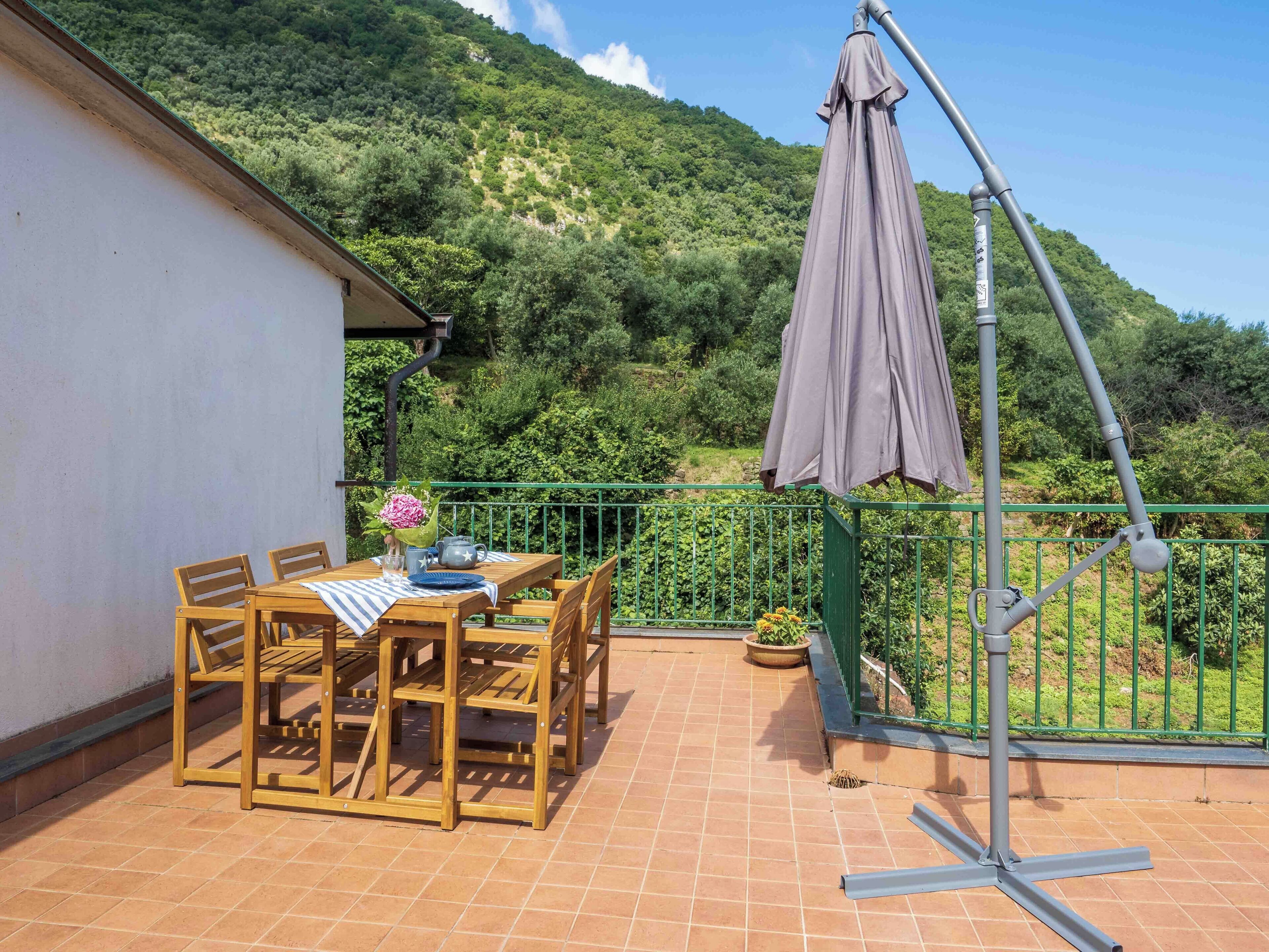 Una mesa de madera con sillas y un mantel en un balcón con barandilla verde, con vistas a una ladera verde y exuberante.
