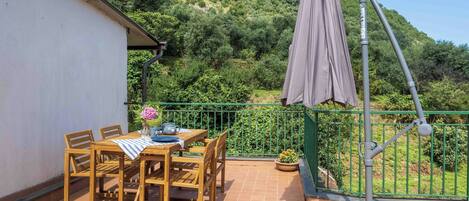 Una mesa de madera con sillas y un mantel en un balcón con barandilla verde, con vistas a una ladera verde y exuberante.