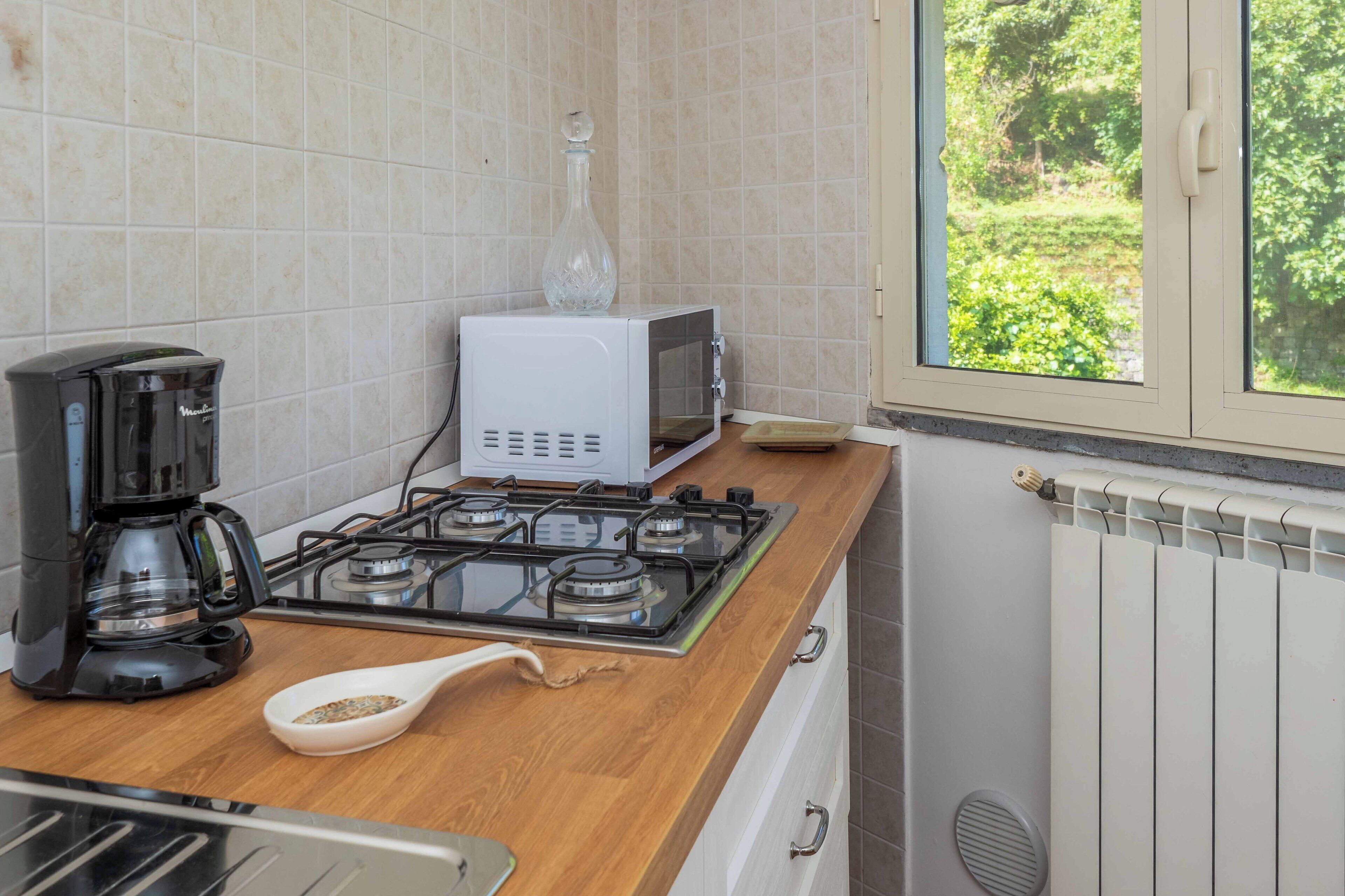A kitchen with a gas stove, a coffee maker, a microwave, and a window with a view of greenery.