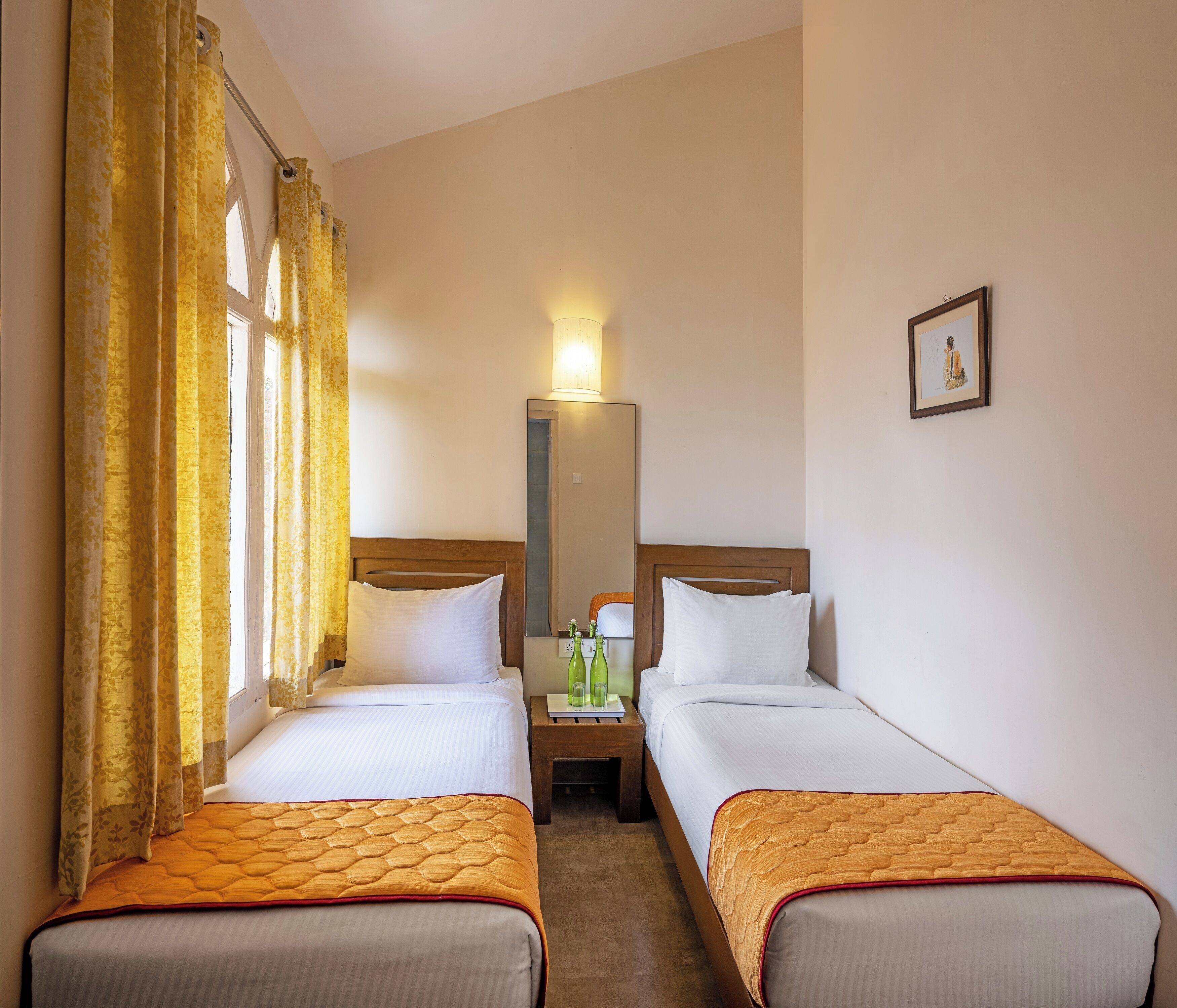 Une chambre d’hôtel avec deux lits simples, une tête de lit en bois, une table de chevet avec une lampe, un miroir et un tableau encadré au mur.
