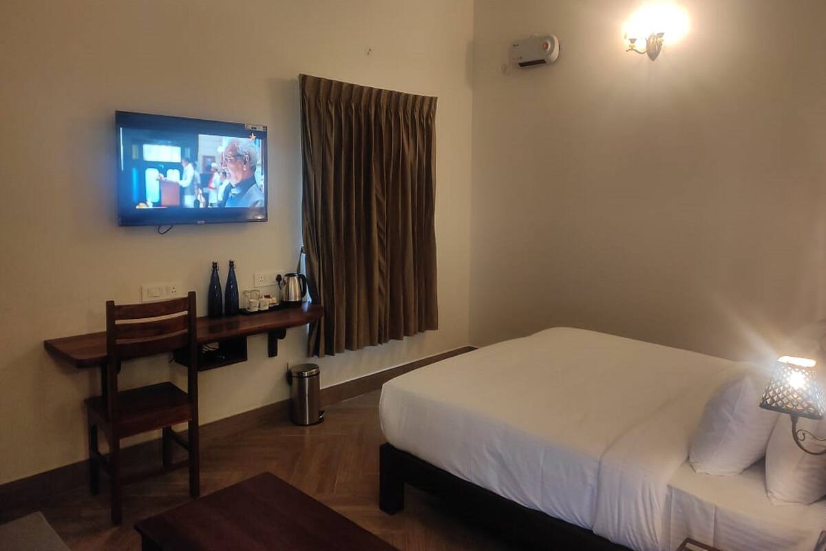 Habitación de hotel con cama, escritorio con silla, televisor montado en la pared, lámpara de noche y ventana con cortinas.