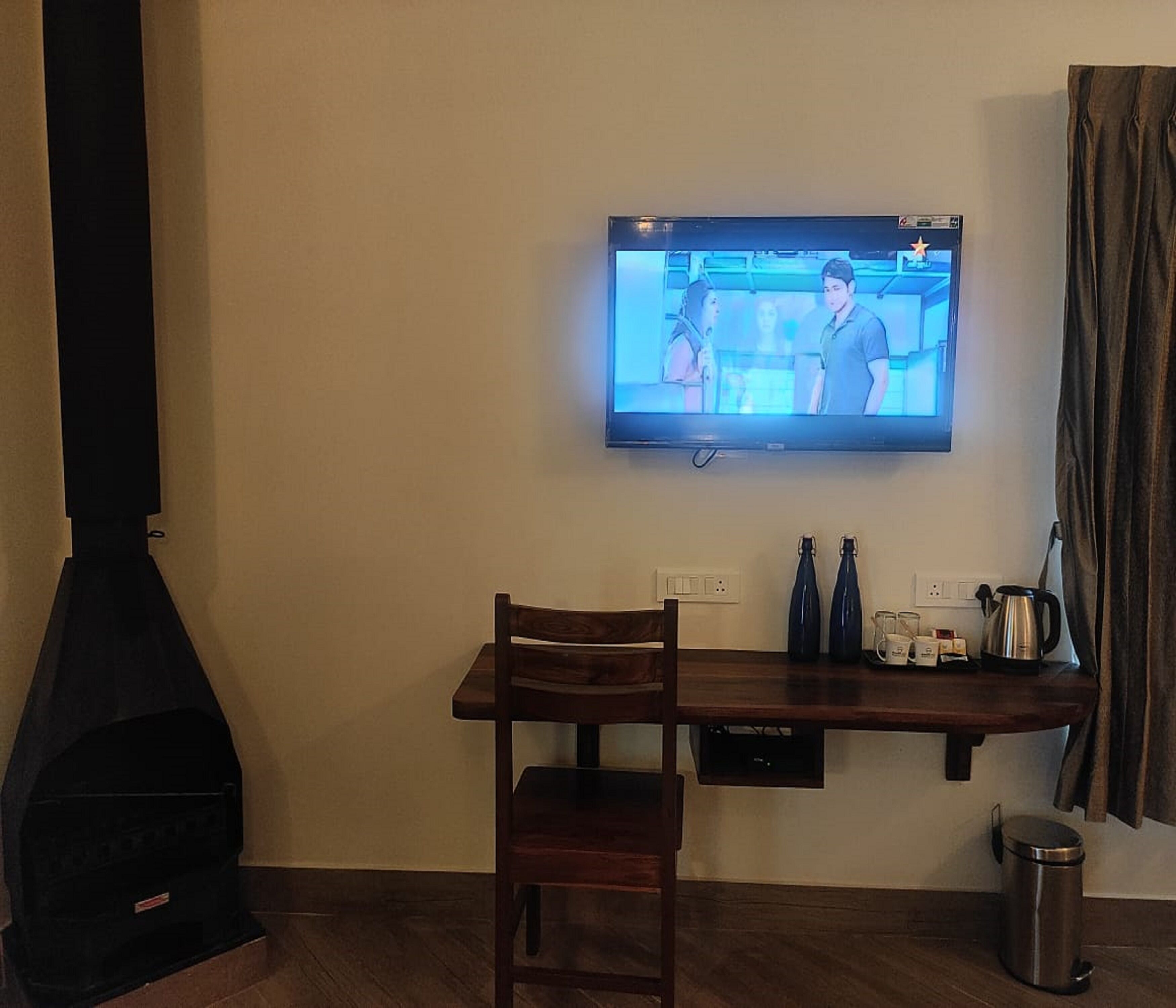 A living room with a TV mounted on the wall, a wooden table, a chair, a fireplace, and a coffee station.