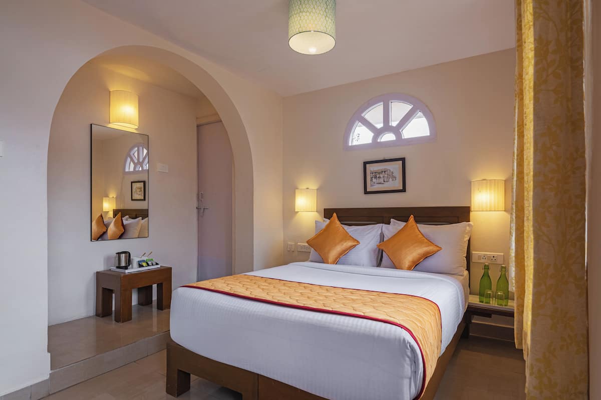 Habitación de hotel con una cama grande, una mesita de noche, un espejo, una ventana y una lámpara de pared.