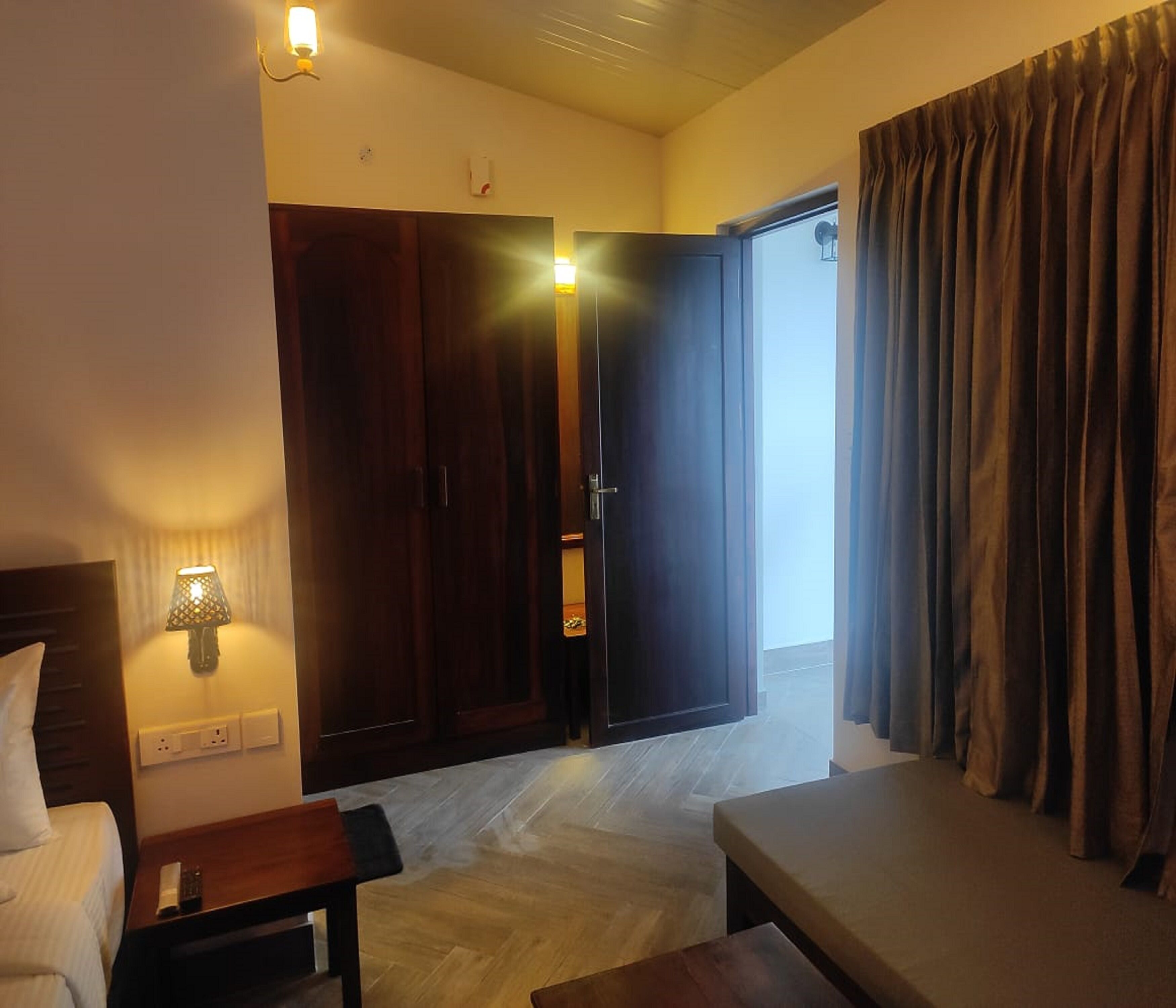 A hotel room with a bed, bedside table, lamp, wardrobe, and a view of the outside.