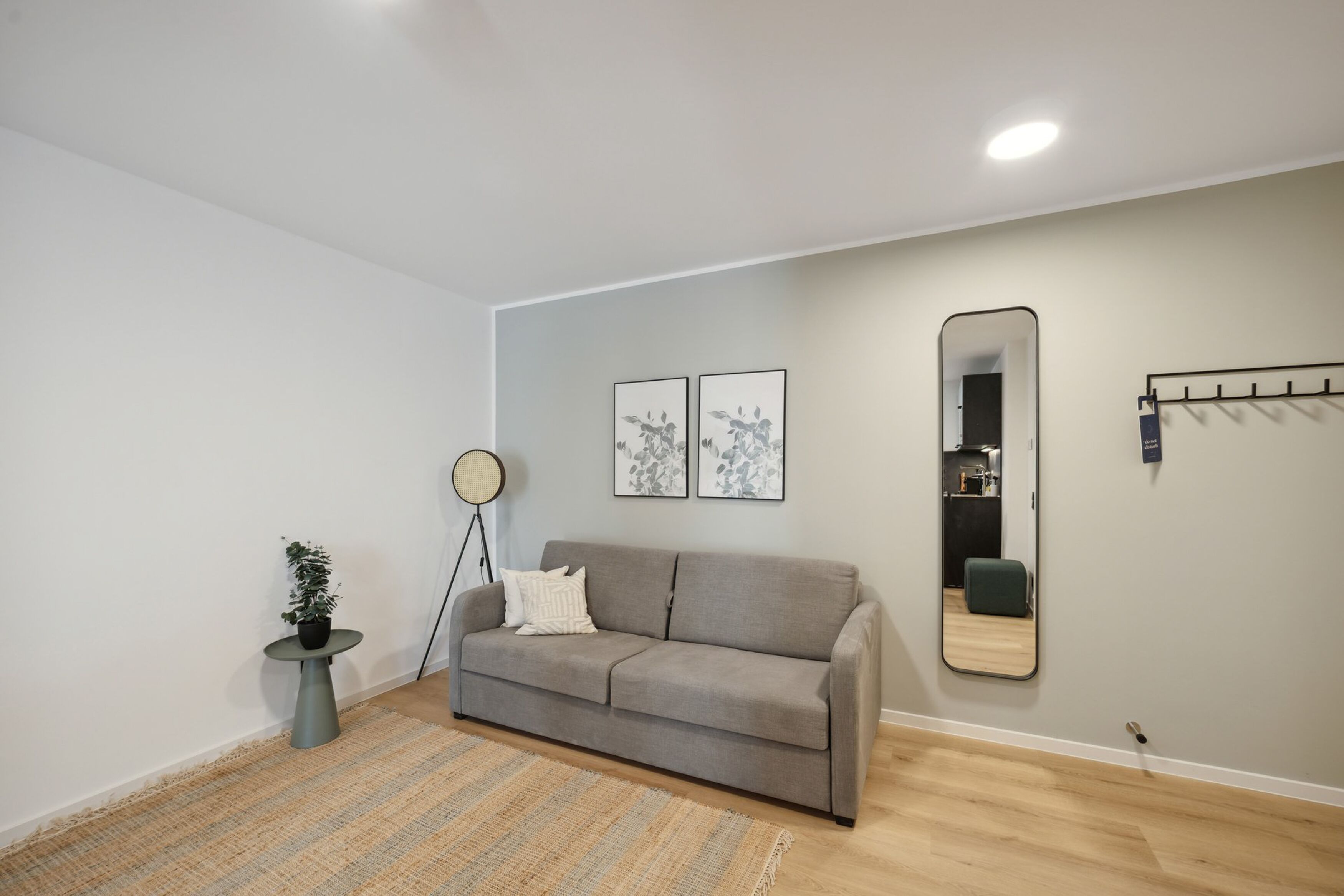 Un salon moderne avec un canapé gris, une lampe sur pied ronde, deux œuvres d’art encadrées, un miroir et une petite plante en pot.