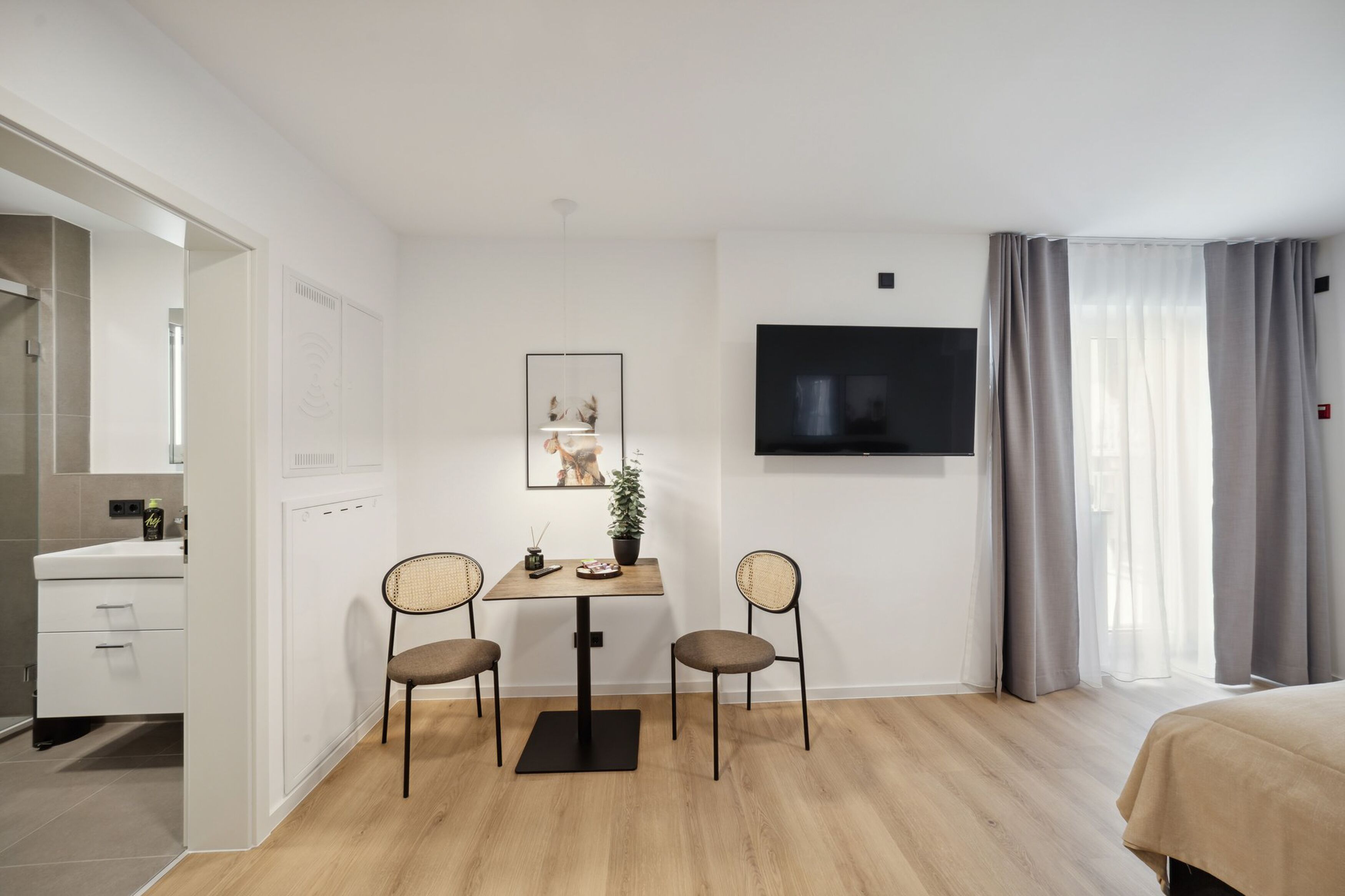 Une chambre d’hôtel moderne avec un plancher en bois, un lit, un téléviseur fixé au mur, une petite table avec deux chaises et une salle de bain visible à travers une porte ouverte.