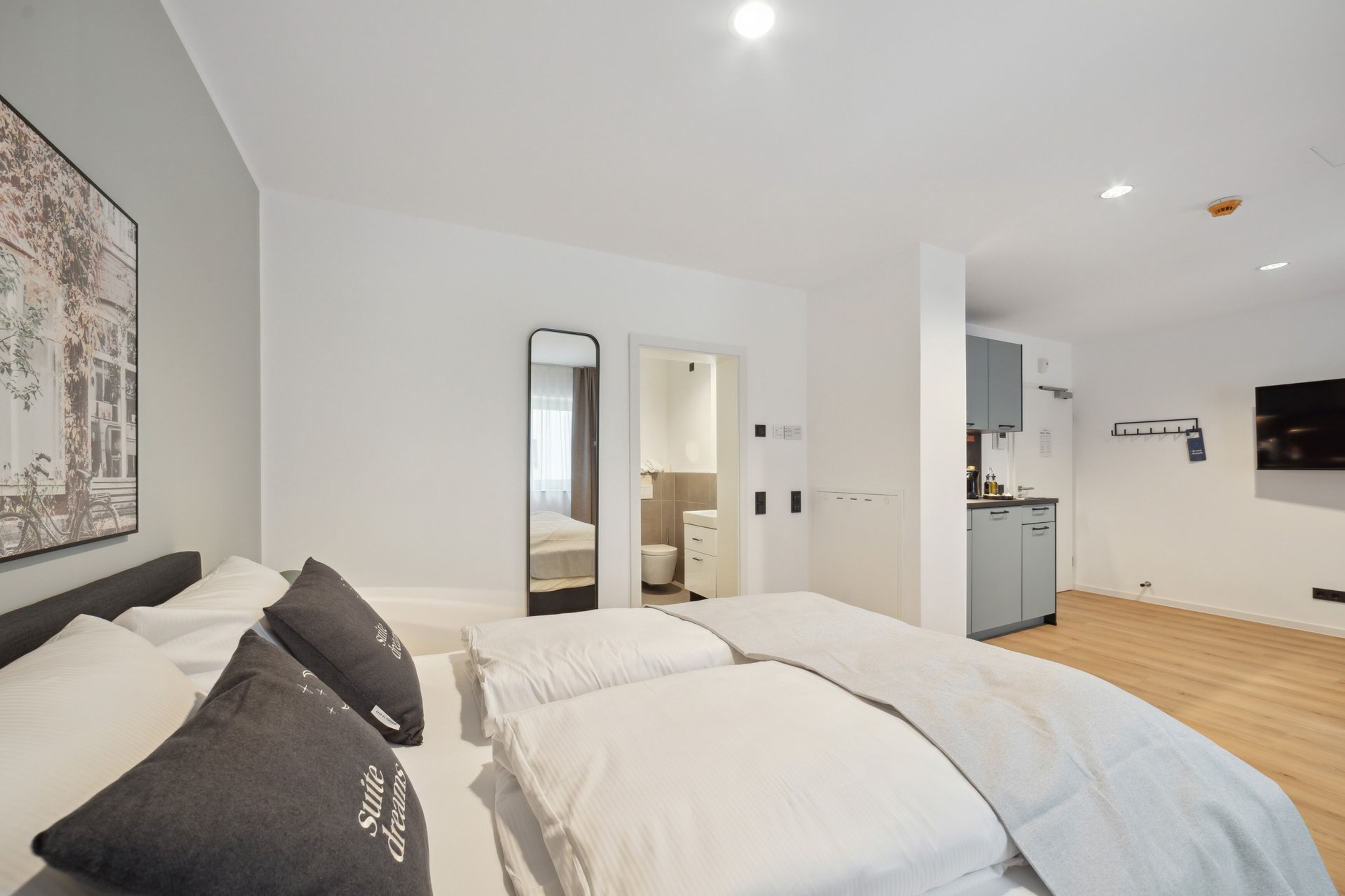 A modern hotel room with a large bed, a small kitchenette, and a bathroom visible through an open door.