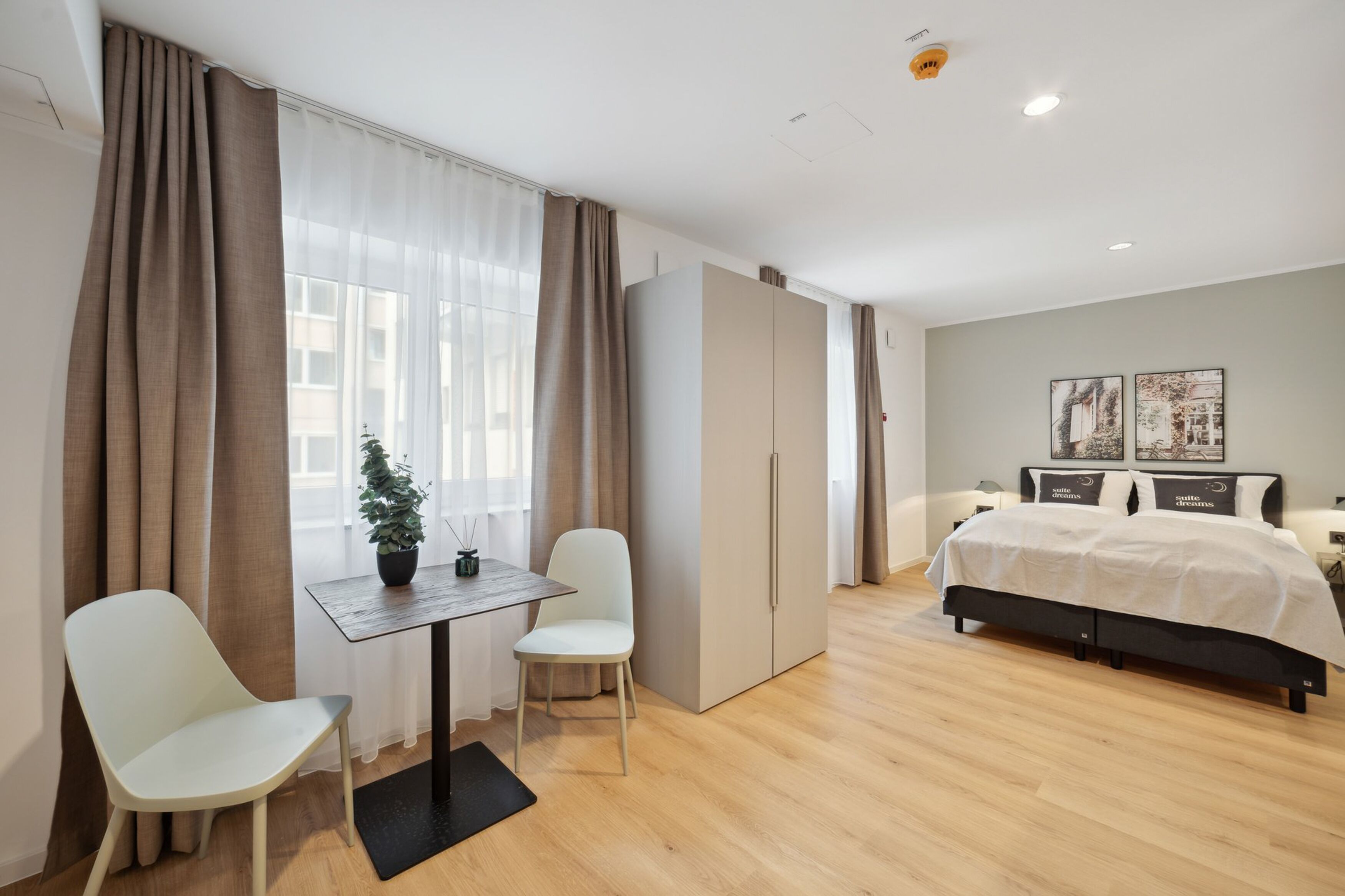Ein modernes Schlafzimmer mit Bett, Schreibtisch, zwei Stühlen, einem Kleiderschrank und einer Zimmerpflanze.