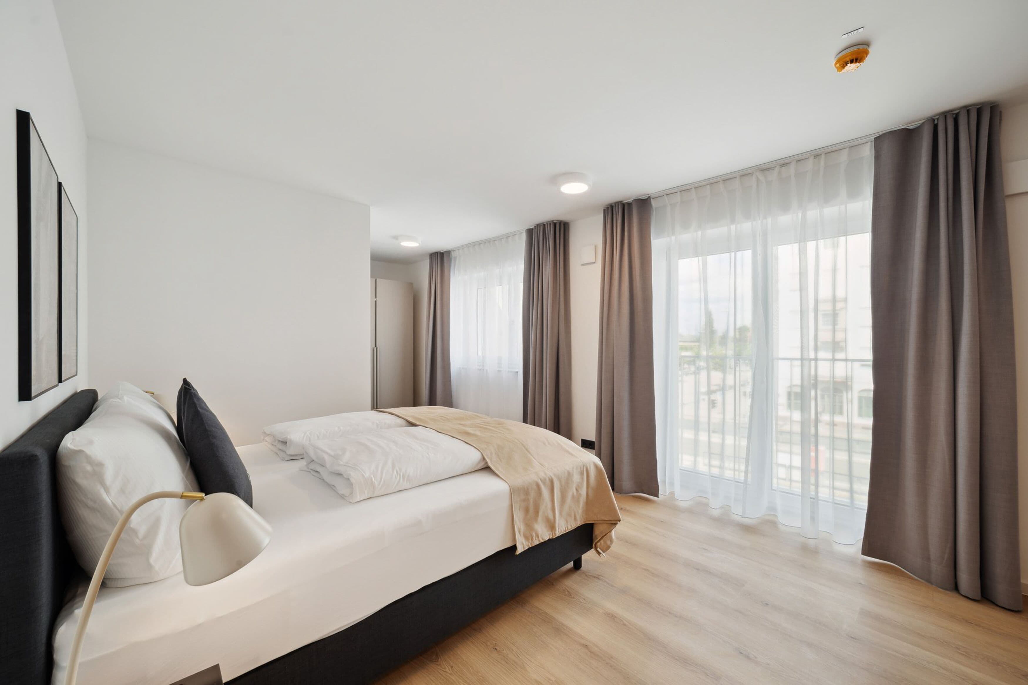Un dormitorio moderno con cama, mesitas de noche y un amplio ventanal con cortinas.
