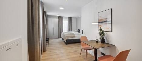 Une chambre d’hôtel moderne avec un plancher en bois, un lit doté d’une tête de lit grise, une petite table avec deux chaises, une plante en pot et un tableau encadré au mur.