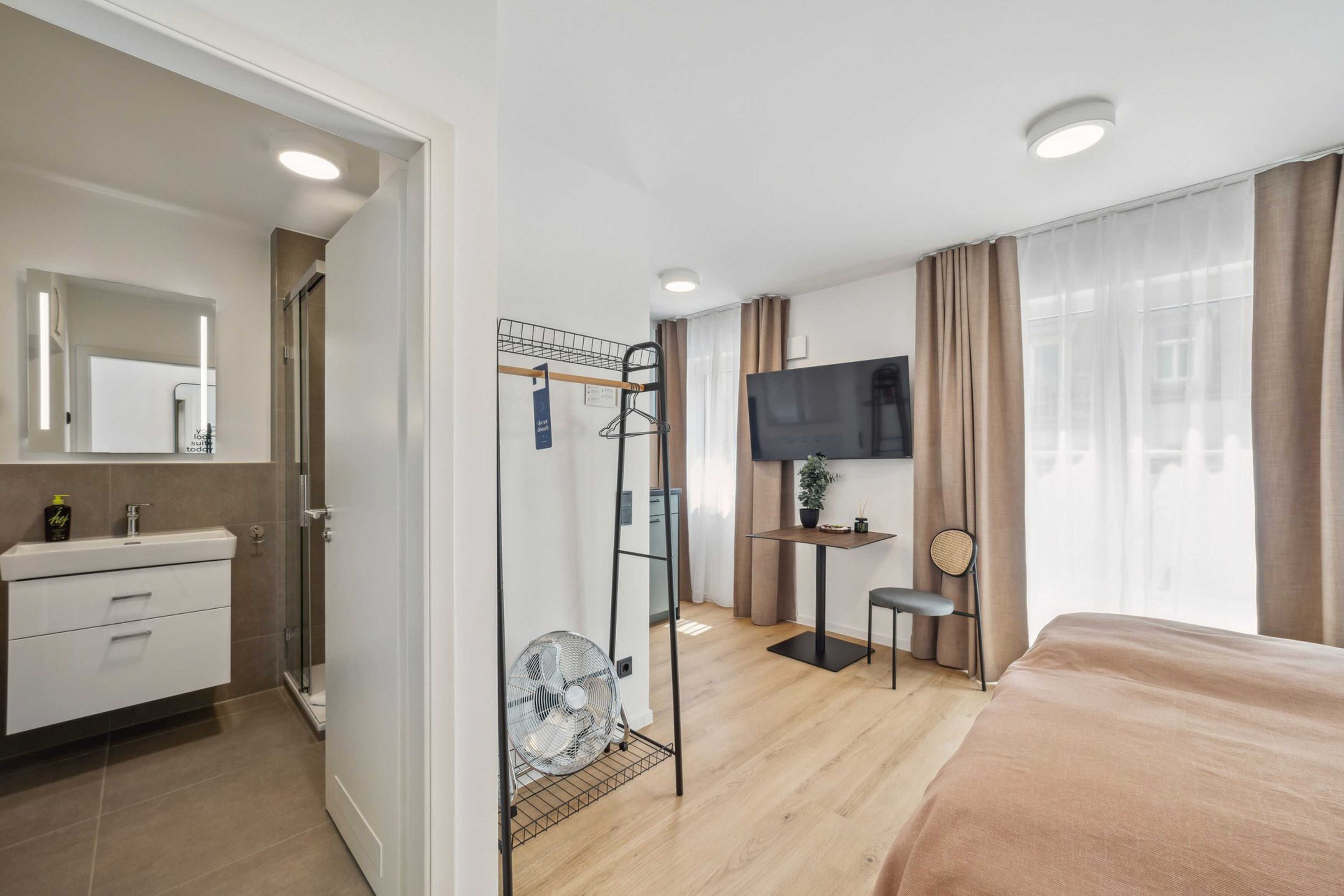 Une chambre d’hôtel comprenant un lit, une télévision, une petite table, une chaise, un ventilateur et une salle de bain avec un miroir et un lavabo.