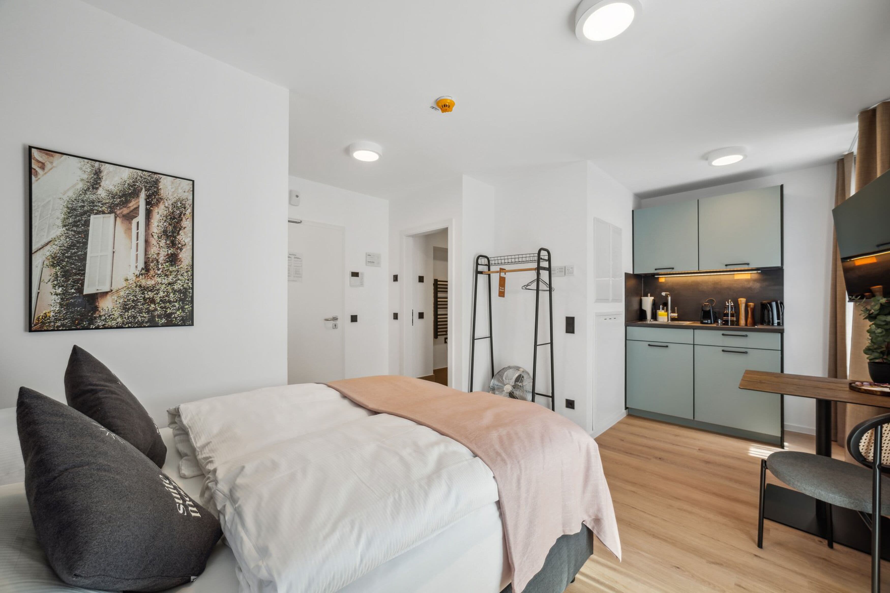 A modern bedroom with a bed, pillows, a bedside table, a desk, a chair, a kitchen area, and a door leading to another room.